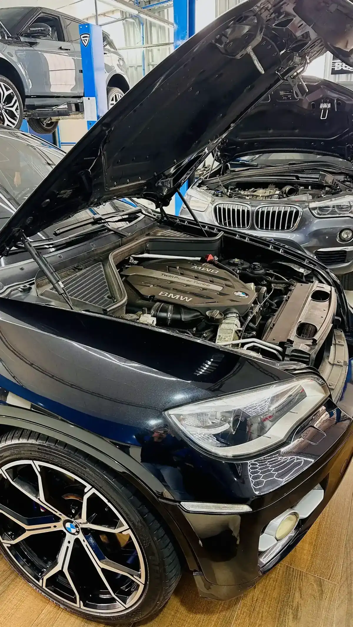 Detalhe do motor de um BMW em close, com outro BMW de capô aberto ao fundo na oficina.