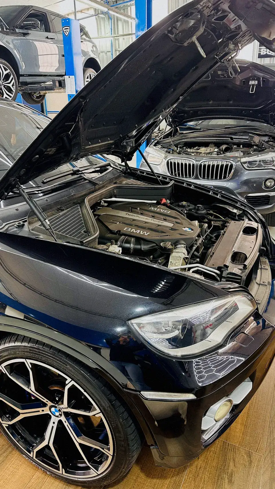 Detalhe do motor de um BMW em close, com outro BMW de capô aberto ao fundo na oficina.