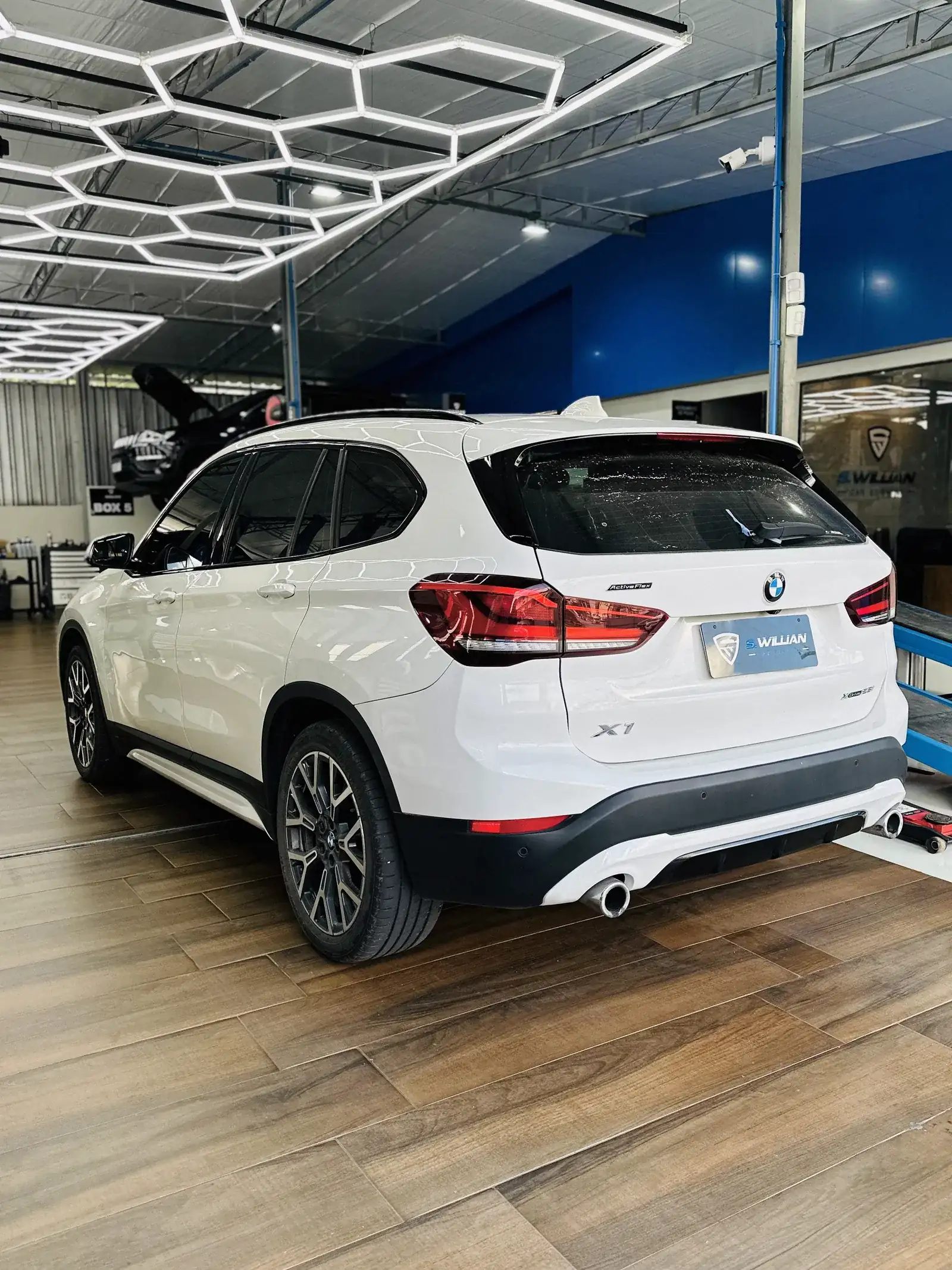 BMW X1 branco visto de trás, sobre piso de madeira, sob teto com iluminação hexagonal em LED.