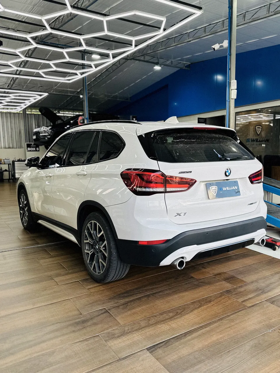 BMW X1 branco visto de trás, sobre piso de madeira, sob teto com iluminação hexagonal em LED.