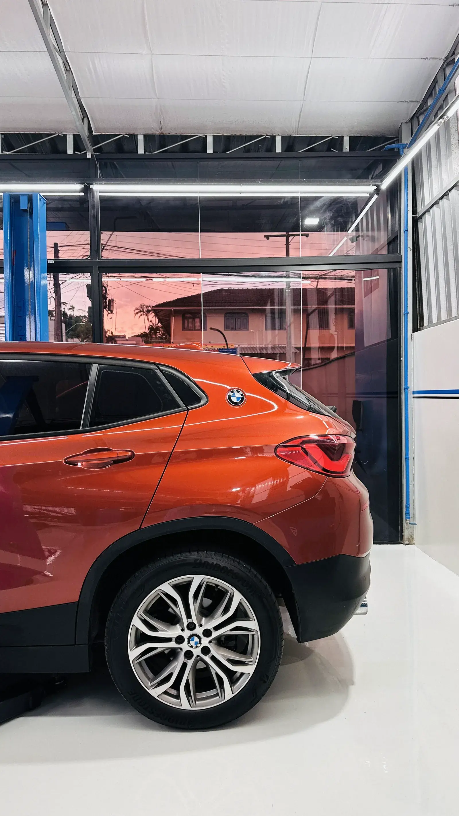 BMW X2 laranja visto de perfil traseiro, com o entardecer refletido no vidro ao fundo da oficina.