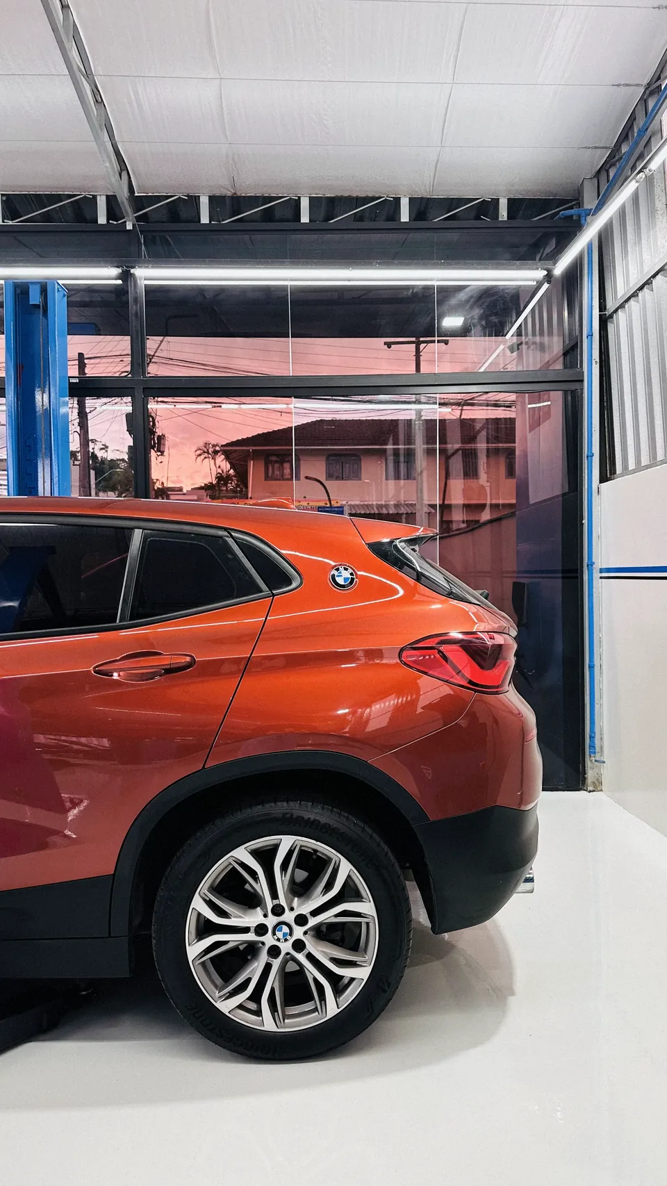 BMW X2 laranja visto de perfil traseiro, com o entardecer refletido no vidro ao fundo da oficina.