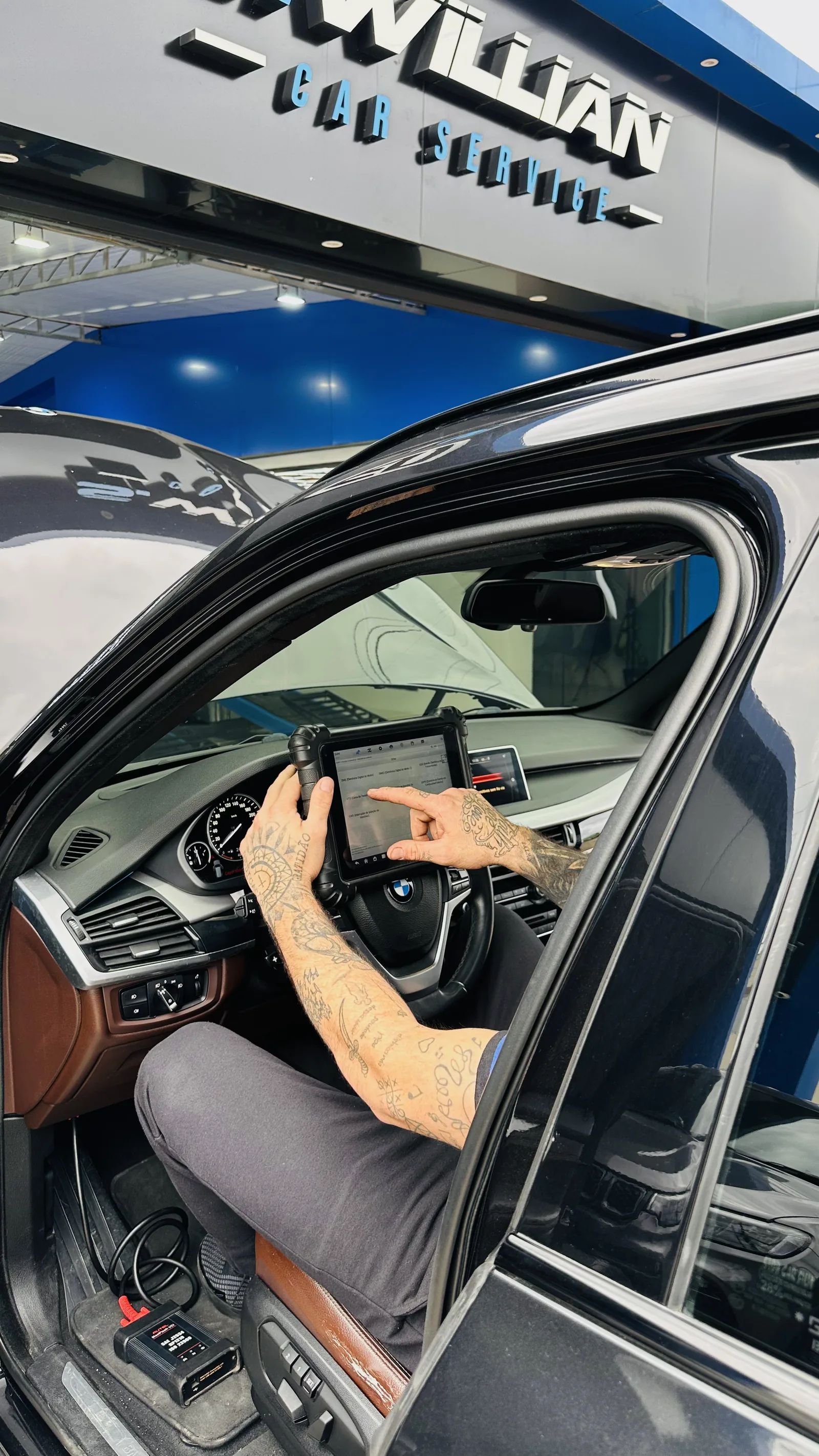 Técnico com o scanner em tablet dentro de um BMW, na frente do letreiro da oficina