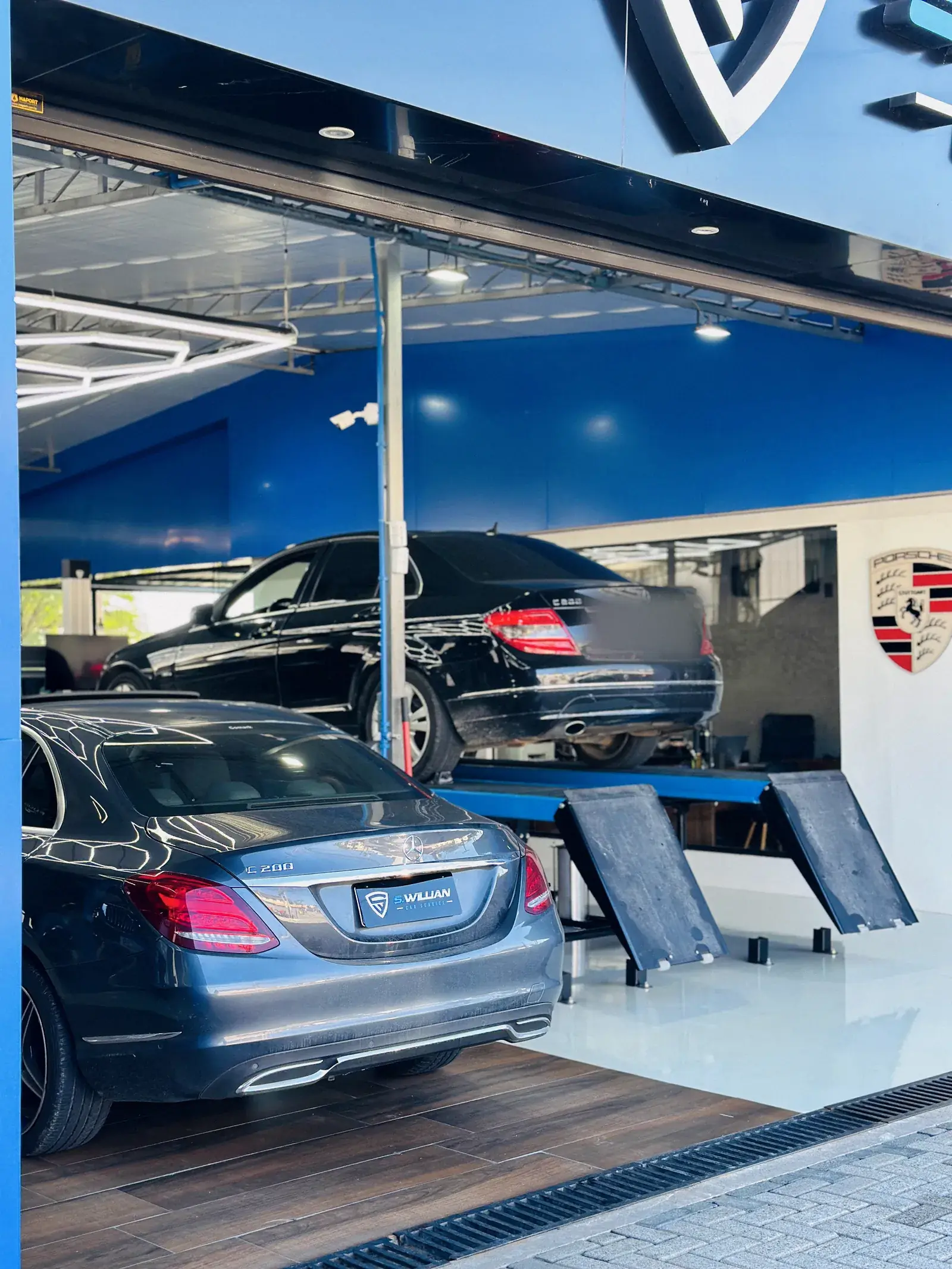 Dois Mercedes-Benz Classe C na fachada da oficina, um em primeiro plano e outro no elevador ao fundo, com o brasão Porsche na parede.