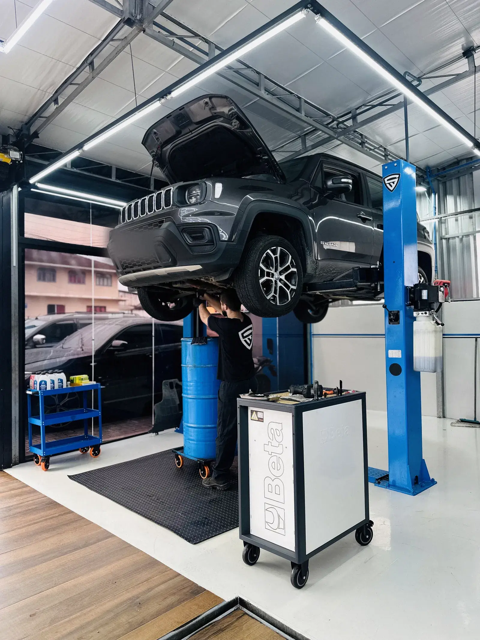 Jeep Renegade no elevador de duas colunas com o capô aberto, técnico dobrado mexendo na suspensão dianteira