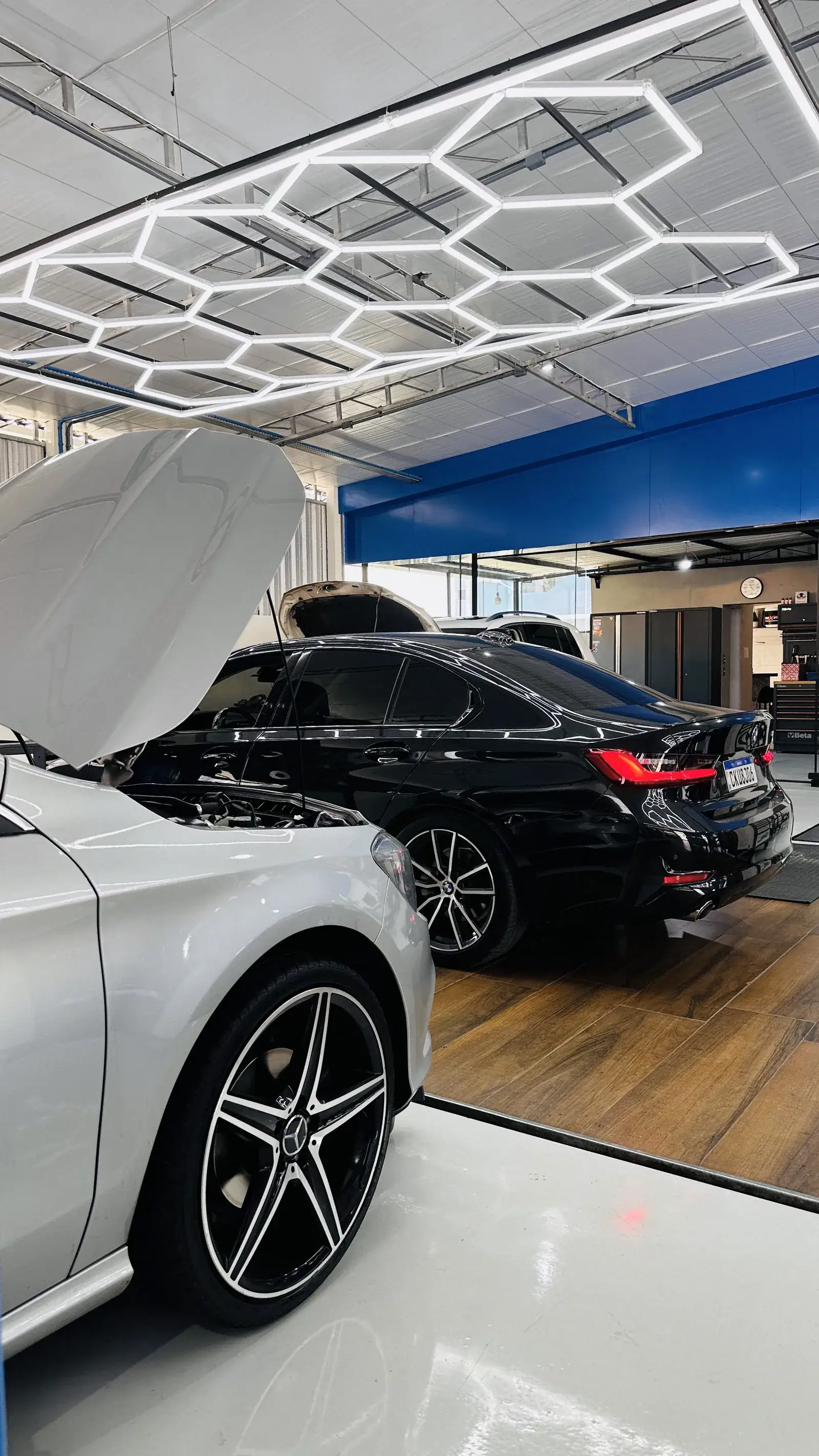 Capô aberto de um Mercedes-Benz com rodas AMG em primeiro plano, BMW desfocado ao fundo, teto hexagonal de LED