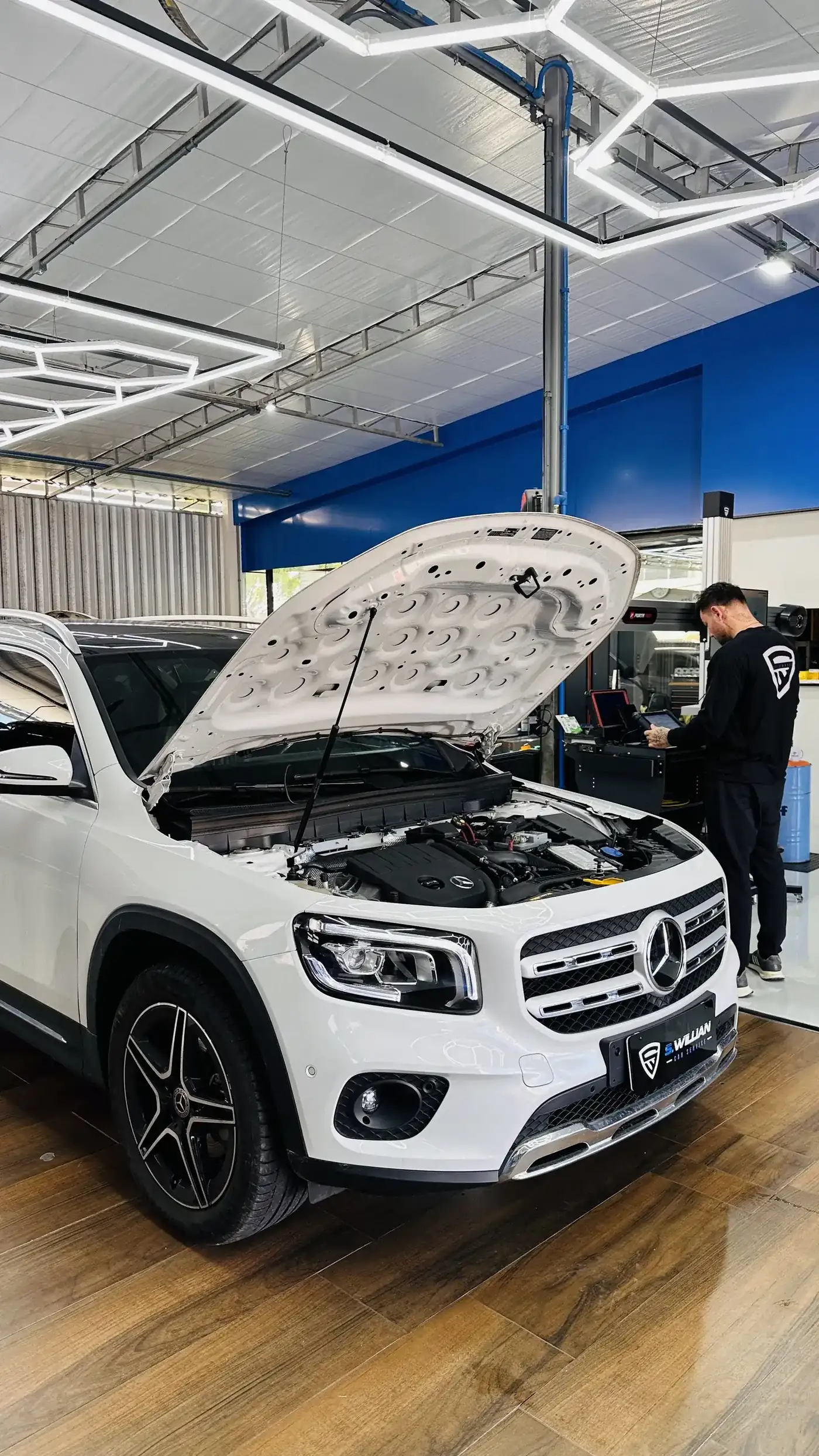 Mercedes-Benz GLB de capô aberto, com técnico trabalhando ao fundo na bancada, sob teto hexagonal de LED.
