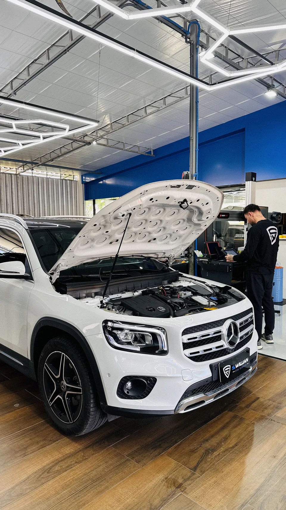 Mercedes-Benz GLB de capô aberto, com técnico trabalhando ao fundo na bancada, sob teto hexagonal de LED.