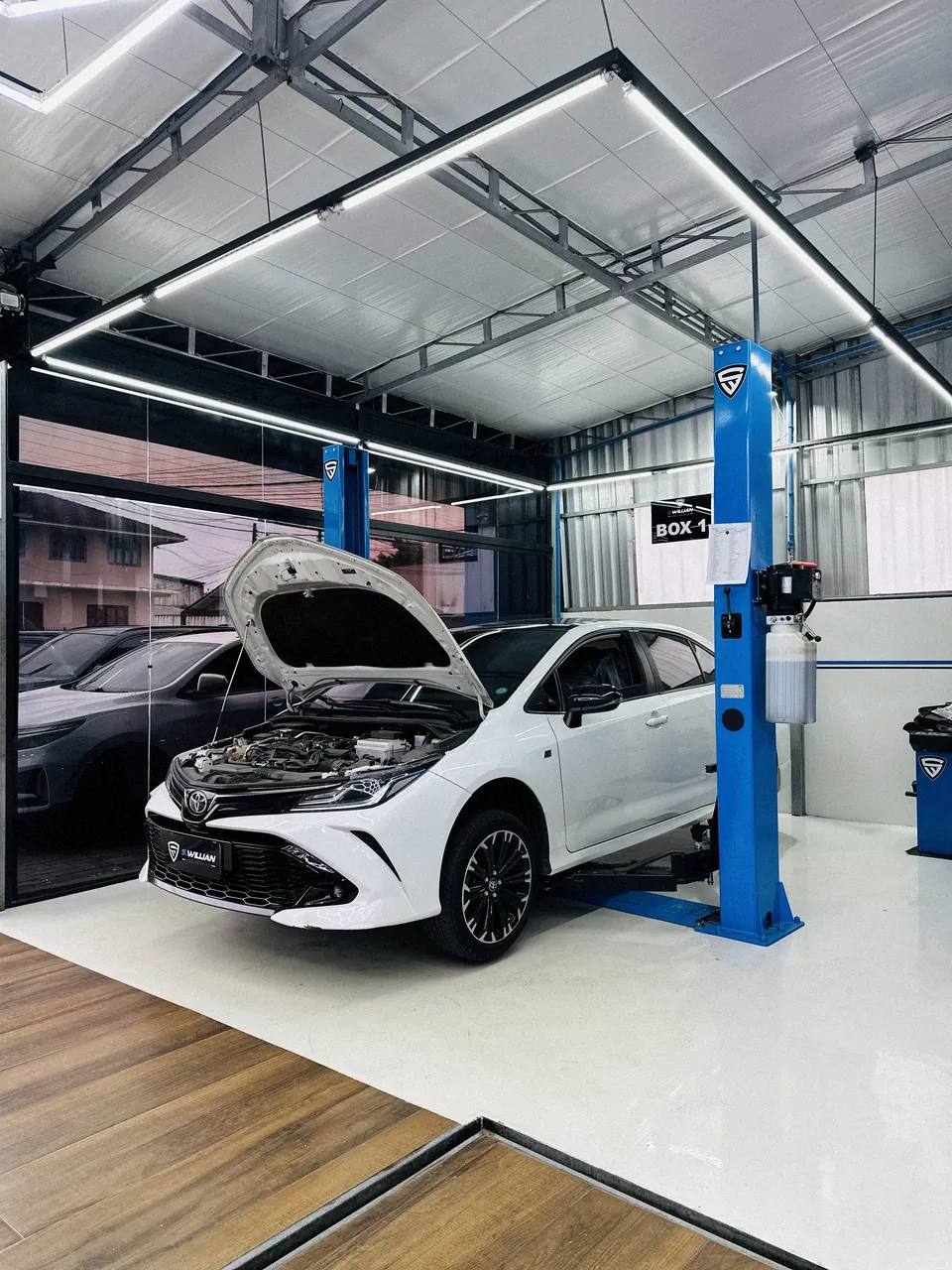 Toyota Corolla com capô aberto, no elevador azul do Box 1, sob teto hexagonal de LED e piso de madeira.