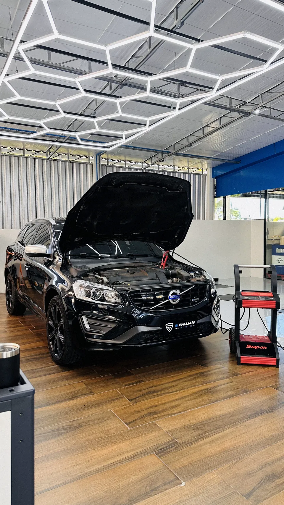 Volvo XC60 com capô aberto, no elevador da oficina, sob teto hexagonal de LED e chão de madeira.