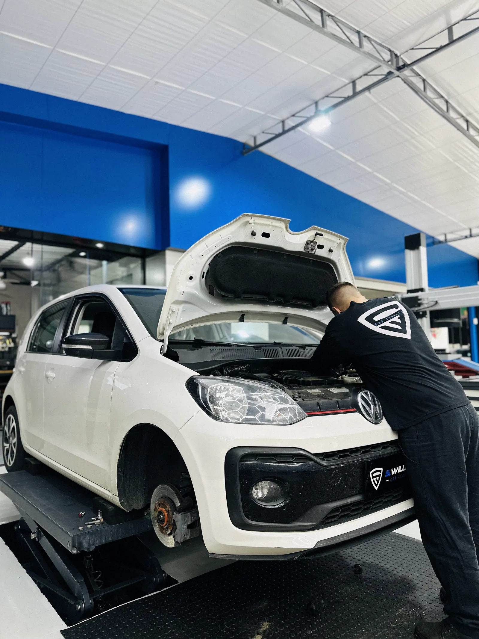 Técnico debruçado sobre o motor de um Volkswagen Up com o capô aberto, parede azul ao fundo